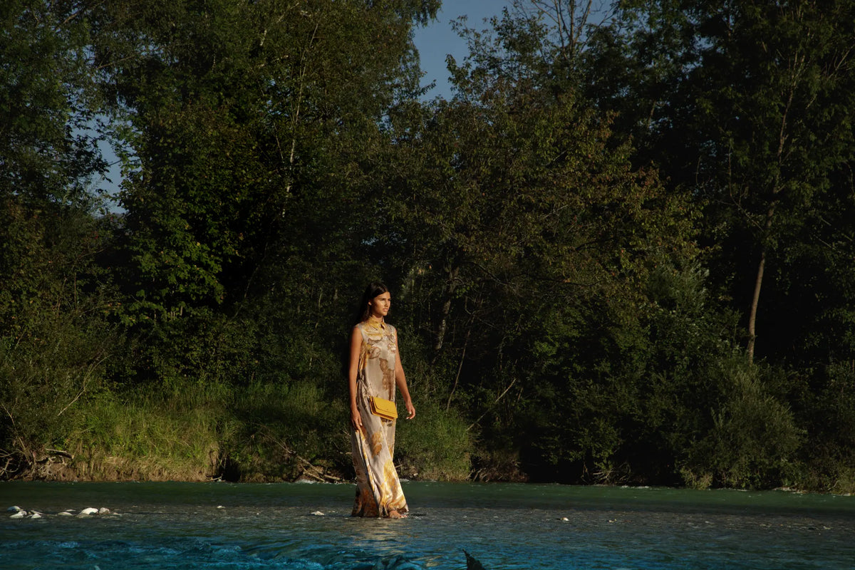 A woman stands in shallow blue water among dense green trees, wearing a long cream and gold patterned dress with a yellow crossbody bag Philini.