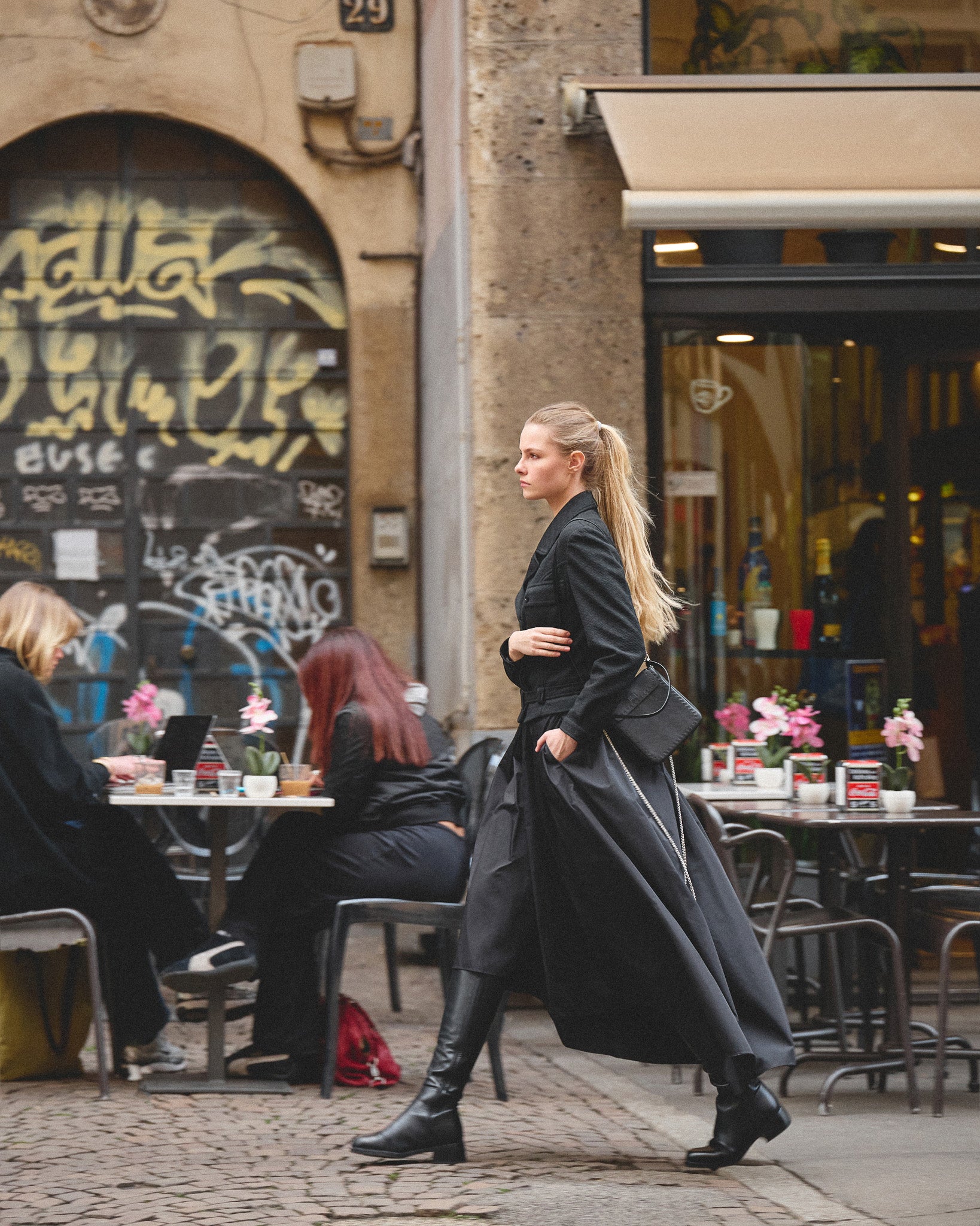 Woman in a long black skirt with philini bags walking past a cafe with tables and people sitting.