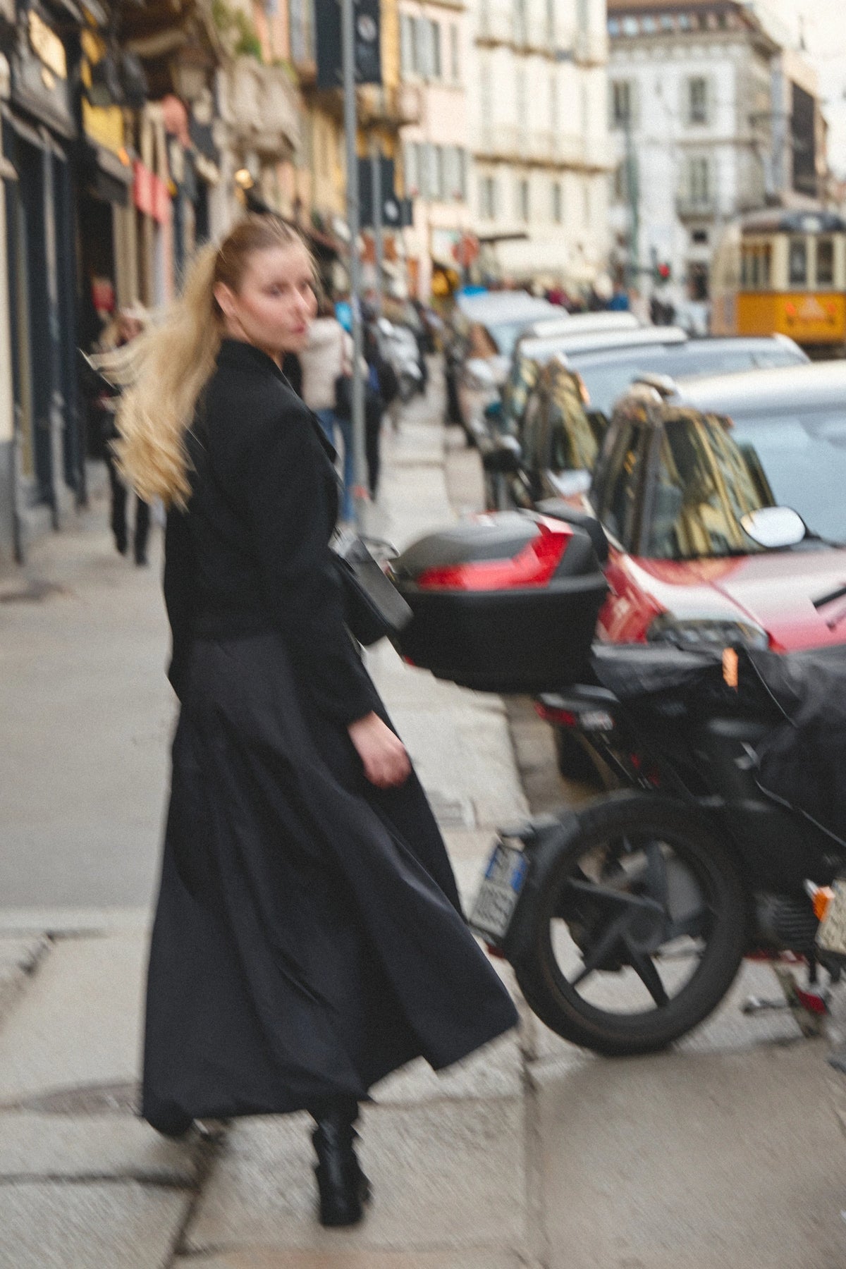 A woman in a long black skirt, a Philini bag and black boots walking on a city street next to parked cars and a motorcycle, photographed with a blurred background.