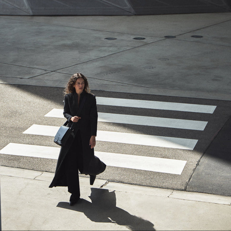 Woman walking on a crosswalk in an urban setting with a modern building in the background at philini campaigns Olga bag black leather