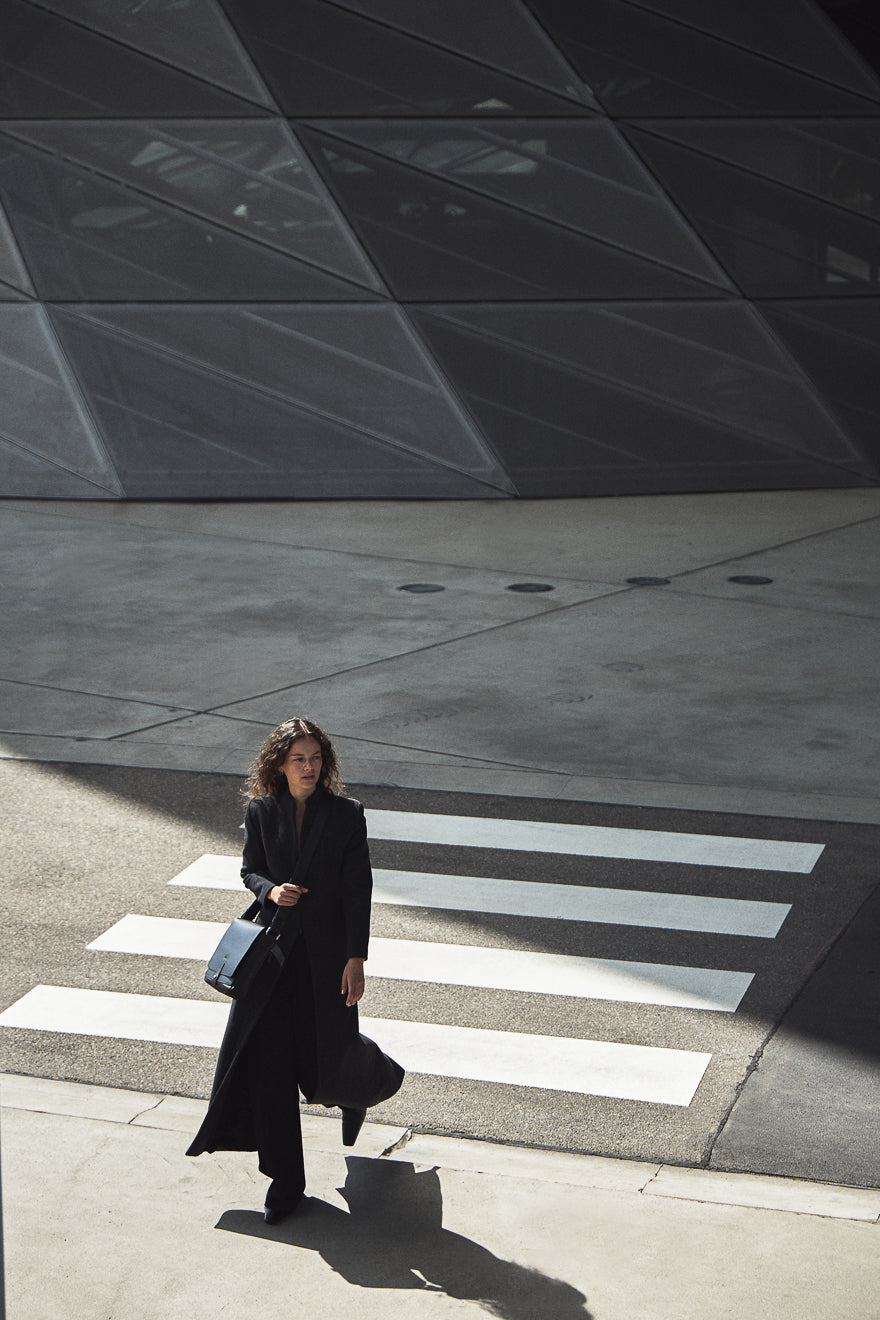 Woman walking on a crosswalk in an urban setting with a modern building in the background at philini campaigns Olga bag black leather